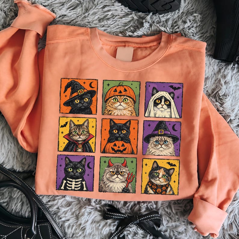 K&ouml;nnte beinhalten: Ein pfirsichfarbenes Sweatshirt mit Rundhalsausschnitt und einem Raster aus neun quadratischen Illustrationen von Katzen in verschiedenen Halloween-Kost&uuml;men, darunter eine Hexe, ein K&uuml;rbis, ein Geist, ein Vampir, ein Skelett und ein Teufel.