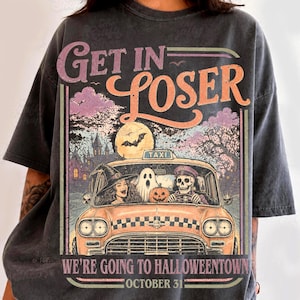 Könnte beinhalten: Ein anthrazitfarbenes T-Shirt mit einem Grafikdruck eines gelben Taxis, in dem eine Hexe, ein Geist und ein Skelett sitzen. Der Text lautet GET IN LOSER WE'RE GOING TO HALLOWEENTOWN OCTOBER 31. Der Hintergrund zeigt ein Spukhaus und einen Vollmond.