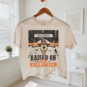 May include: A cream-colored short-sleeve t-shirt featuring a graphic print of a bull skull wearing a witch hat. The design includes the text HALLOWEEN, RAISED ON 90s HALLOWEEN, silhouettes of bats, a raven, and bare tree branches in black and orange.