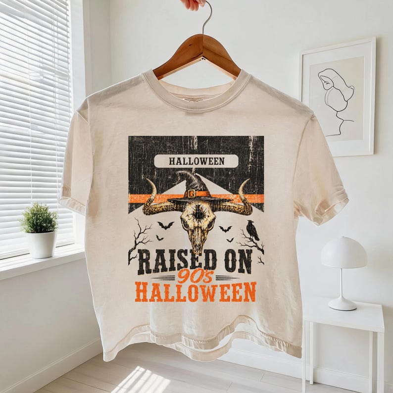 May include: A cream-colored short-sleeve t-shirt featuring a graphic print of a bull skull wearing a witch hat. The design includes the text HALLOWEEN, RAISED ON 90s HALLOWEEN, silhouettes of bats, a raven, and bare tree branches in black and orange.