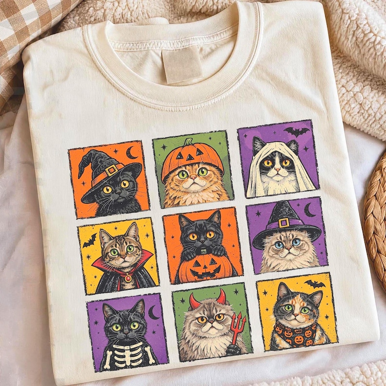 K&ouml;nnte beinhalten: Ein cremefarbenes T-Shirt mit einem Raster aus neun quadratischen Illustrationen von Katzen in verschiedenen Halloween-Kost&uuml;men, darunter eine Hexe, ein K&uuml;rbis, ein Geist, ein Vampir, ein Teufel und ein Skelett, vor orangefarbenen, gr&uuml;nen, violetten und gelben Hintergr&uuml;nden.