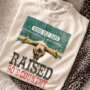 Könnte beinhalten: Ein cremefarbenes T-Shirt mit Rundhalsausschnitt und einer Grafik eines Longhorn-Schädels auf einem verwaschenen grünen rechteckigen Hintergrund. Der Text GOOD OLD DAYS, RAISED und 90'S COUNTRY ist in schwarzer und roter Schrift.