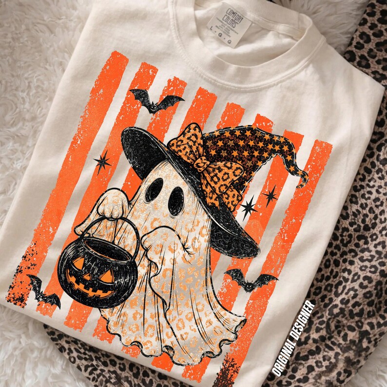 May include: A cream-coloured t-shirt featuring a graphic of a ghost wearing a witch hat with a leopard print pattern. The ghost holds a black jack-o'-lantern bucket. The background of the graphic consists of vertical orange stripes, small black bats, and star shapes. The shirt has a Comfort Colors label.