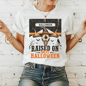 May include: A white short-sleeve t-shirt featuring a graphic of a bull skull wearing a witch hat. The design includes the text HALLOWEEN, RAISED ON 90's HALLOWEEN, and illustrations of bats and a raven against a black and orange background.