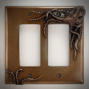May include: A double light switch cover with a bronze finish features a 3D sculpted octopus design. The octopus is depicted with tentacles wrapping around the switch plate.