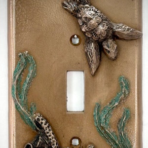 May include: A light brown ceramic light switch cover with a 3D design of a seahorse, coral, and seaweed. The seahorse is brown and has a textured surface. The coral is brown and has a textured surface. The seaweed is green and has a textured surface.