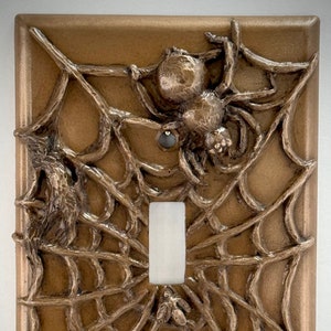 Könnte beinhalten: Ein brauner Lichtschalter mit einem 3D-Spinnennetz-Design. Eine Spinne befindet sich in der Mitte des Netzes, und eine Fliege ist im Netz unter dem Schalter gefangen.