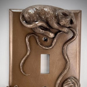 May include: A brown light switch cover with a sculpted design of two cats playing with a ball of yarn. The top cat is sleeping on the yarn, while the bottom cat is pouncing on the yarn.