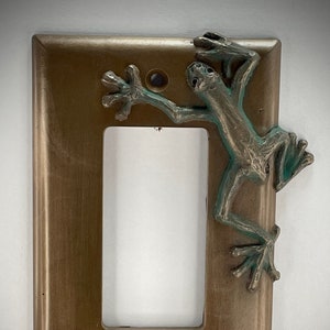 May include: A bronze light switch cover with a green frog sculpture attached to the right side.
