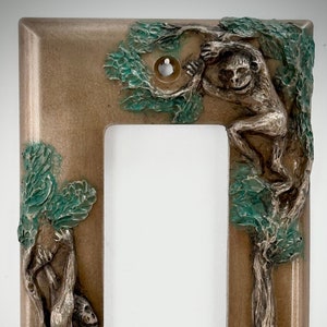 May include: A light brown wall plate with a decorative design of two monkeys swinging from a tree. The tree is painted in shades of green and brown. The plate is designed to cover a light switch.