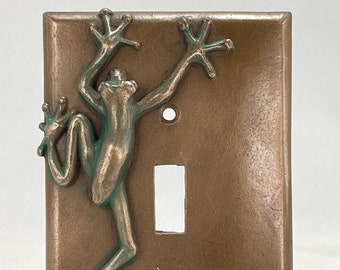 Puede incluir: Una tapa de interruptor de luz de bronce con una escultura de rana verde unida. La rana está subiendo por la tapa con los brazos y las piernas extendidos.
