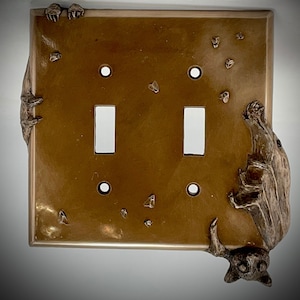 May include: A brown metal light switch cover with two toggle switches and two bats on either side. The bats are sculpted in a 3D design and appear to be climbing the switch cover.
