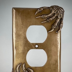 May include: A gold-colored outlet cover with two sculpted dinosaur claws on either side. The claws are detailed with scales and sharp points.