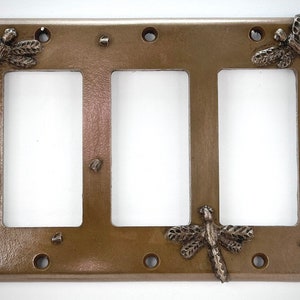 May include: A triple light switch cover plate with a bronze finish and three dragonfly designs. The dragonflies are in a silver finish and have detailed wings.