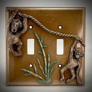May include: A brown ceramic light switch cover with two toggle switches. The cover features a 3D design of two monkeys swinging on a rope with bamboo plants in the background.