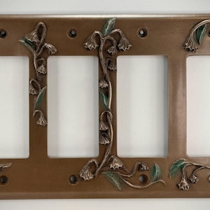 May include: A decorative light switch plate with four openings. The plate is bronze-colored with an embossed floral design featuring bell-shaped flowers and green leaves. The plate has four screw holes.