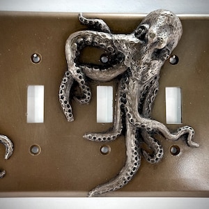 May include: A brown light switch cover with a three-dimensional silver octopus design. The octopus has tentacles that wrap around the switch cover and extend to the sides.