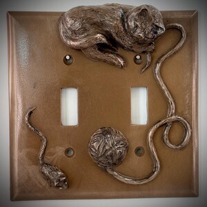 May include: A brown double light switch cover with a 3D sculpted design of a cat sleeping on a ball of yarn. The cat is holding a string in its paw and a mouse is at the bottom of the switch cover.