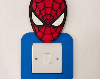 May include: A red and black Spider-Man mask decorates a blue light switch cover. The switch is white, and the mask has white eye shapes. The design is mounted on a white wall.