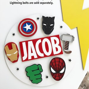 May include: A white circular name plaque with the name "JACOB" in large white letters on a red background. The plaque features superhero-themed cutouts, including Captain America's shield, Iron Man's mask, and Spider-Man's mask.