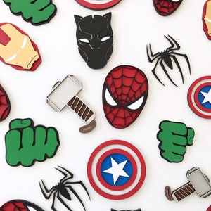 May include: A collection of colourful wooden cutouts featuring superhero designs. Includes Spider-Man, Captain America's shield, Hulk's fist, Thor's hammer, Iron Man's mask, Black Panther's mask, and a black spider. The cutouts are on a white background.