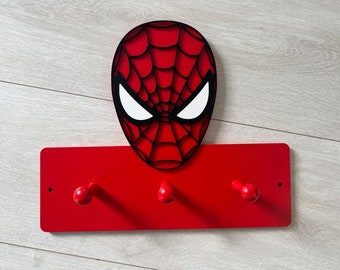 May include: A red wall-mounted coat rack featuring the face of Spider-Man. The rack has three red knobs for hanging items. The Spider-Man face is red with black webbing and white eye details, creating a 3D effect.