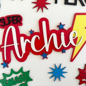 May include: A custom wooden wall sign featuring the name Archie in white script lettering layered over a red background. The design includes a yellow lightning bolt, blue star accents, and comic book style speech bubbles with the words Super and Smash.