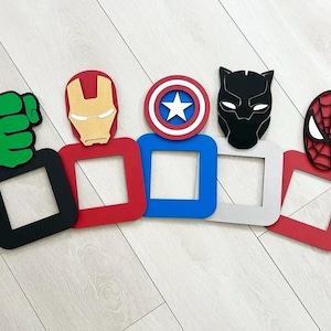 May include: Superhero-themed wall decorations featuring characters like Iron Man, Captain America, Black Panther, and Spider-Man. Each character is represented with a mask or symbol, paired with a square frame in colours such as red, blue, and black. The Hulk's fist is also included.