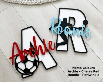May include: Wooden name cutouts for kids, featuring the names "Archie" and "Ronnie". The letter "A" has a cherry red "Archie" with a soccer ball. The letter "R" has a periwinkle "Ronnie" with a soccer player silhouette. The letters are white with black borders.