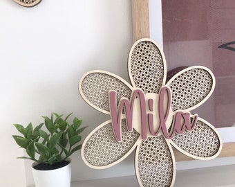 May include: A wooden flower-shaped wall decor with a woven center and the name "Mila" in pink script. The flower is displayed on a white shelf, with a small potted plant to the left and a framed picture in the background.