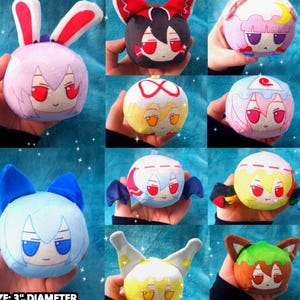 May include: A collection of nine plush character dolls, each approximately 3 inches in diameter, with unique facial expressions and accessories like bunny ears, bows, and wings. The dolls are in various colors, including pink, blue, yellow, and brown. The text "SIZE: 3" DIAMETER COMES WITH BEAD CHAIN" is at the bottom.