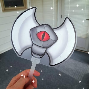 May include: A cartoon-style hand fan shaped like a battle axe. The fan features a white blade with a gray handle and a central design of a gray stone with a red eye. The fan is held by a clear plastic handle.