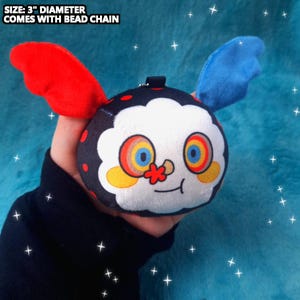May include: A plush toy with a round black body, red and blue wings, and a white face with colorful eyes and a flower nose. The toy is 3" in diameter and comes with a bead chain.