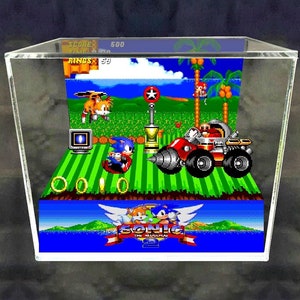 Könnte beinhalten: Ein 3D-Diorama einer Szene aus dem Videospiel Sonic the Hedgehog 2. Das Diorama zeigt Sonic, Tails und Dr. Robotnik in einem grünen Feld mit blauem Himmel. Das Diorama ist in einem durchsichtigen Acryl-Würfel eingeschlossen. Der Text "SONIC THE HEDGEHOG 2" ist am unteren Rand des Dioramas sichtbar.