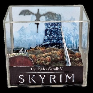 May include: A clear acrylic cube display case with a 3D diorama of a snowy mountain landscape from the video game "The Elder Scrolls V: Skyrim". The diorama features a dragon flying over a tower, a sword, and a snowy mountain range in the background. The base of the display case has the game's logo, "The Elder Scrolls V: Skyrim".