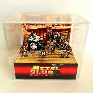 Può includere: Un diorama 3D di una scena del videogioco Metal Slug X. Il diorama presenta un carro armato pixelato, soldati e un edificio. Il diorama è in una teca di acrilico trasparente. Il testo "METAL SLUG X SUPER VEHICLE-001" è stampato sulla base del diorama.