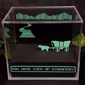 Peut inclure: Un cube de présentation en acrylique transparent contenant une plateforme noire avec un chariot pixélisé vert clair tiré par un bœuf. L'arrière-plan présente un paysage pixélisé vert et le panneau avant affiche le texte YOU HAVE DIED OF DYSENTERY.