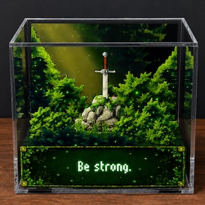 Può includere: Diorama in pixel art in un cubo di acrilico trasparente. La scena raffigura una spada nella roccia circondata da alberi e fogliame verdi. Il testo "Be strong." è visualizzato alla base del cubo.