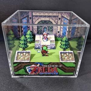 May include: A 3D diorama of The Legend of Zelda: Ocarina of Time encased in a clear acrylic cube. The scene features pixelated characters, green trees, and flower beds on a grassy base, with a stone temple backdrop and the game title printed.