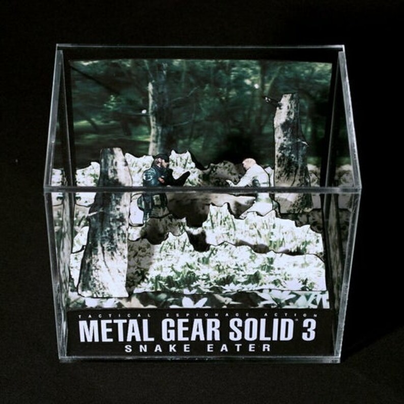 Puede incluir: Un diorama de una escena del videojuego Metal Gear Solid 3: Snake Eater. El diorama presenta dos figuras en un entorno de jungla. El texto "Metal Gear Solid 3: Snake Eater" es visible en la base del diorama.