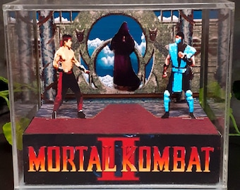 Op de afbeelding: Een Mortal Kombat II diorama in een doorzichtige acryl doos. De scène toont twee gepixelde videogamepersonages die tegenover elkaar staan op een rood platform, met een donkere figuur met kap in een stenen boog op de achtergrond.