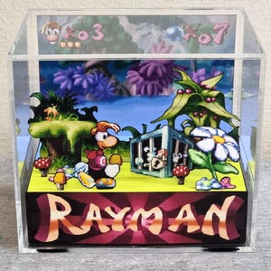 May include: A clear acrylic display box featuring a 3D diorama of the video game Rayman. The scene includes the Rayman character, a cage, flowers, and the game's title. The background shows a colorful landscape with the numbers 3 and 7.