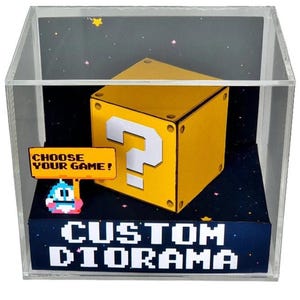 May include: A clear acrylic display box containing a yellow question mark block, a small pixelated character holding a sign that reads CHOOSE YOUR GAME!, and a base with the text CUSTOM DIORAMA against a dark background with colorful stars.