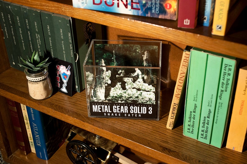 Puede incluir: Una caja de presentaci&oacute;n de cubo de pl&aacute;stico transparente con una imagen en blanco y negro de una escena de bosque en su interior. La caja tiene una etiqueta negra en la parte inferior que dice "METAL GEAR SOLID 3 SNAKE EATER".
