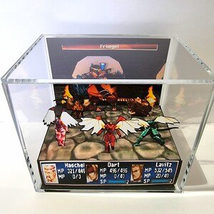 Pode incluir: Um diorama 3D de uma cena de batalha de um videogame com quatro personagens: Haschel, Dart, Lavitz e Fruegel. Os personagens est&atilde;o em p&eacute; em um terreno rochoso marrom com chamas ao fundo. O diorama est&aacute; fechado em uma caixa de acr&iacute;lico transparente.