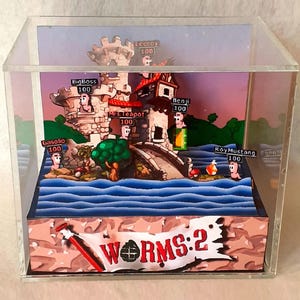 Op de afbeelding: Een 3D-diorama van het videospel Worms 2 in een doorzichtige acrylblok. De scène toont een stenen kasteel, een brug en water, met verschillende wormpersonages genaamd BigBoss, Mrs.Teapot, Benji, Leeroy, Gasolio en RoyMustang, elk met 100 levenspunten.