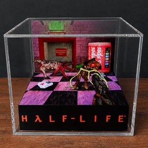 Puede incluir: Una caja de exhibición de acrílico transparente que contiene un diorama de Half-Life. La escena presenta un suelo a cuadros, una máquina de refrescos roja, una puerta restringida y dos criaturas alienígenas. Un arma negra está al frente.