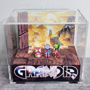Puede incluir: Una caja de exhibición de acrílico transparente que contiene un diorama 3D del videojuego Grandia. Dentro, tres pequeñas figuras de personajes se encuentran sobre una plataforma con textura de madera frente a un fondo de paisaje impreso.