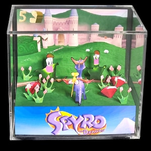 Puede incluir: Un diorama en cubo de acrílico transparente que muestra una escena en 3D del videojuego Spyro the Dragon. La pieza incluye una figura de dragón morado, césped verde, gemas rojas y un fondo de castillo. El logo aparece en el panel frontal.
