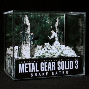 Puede incluir: Una vitrina de pl&aacute;stico transparente con un diorama en su interior que representa una escena del videojuego Metal Gear Solid 3: Snake Eater. El diorama presenta dos figuras, una con un traje de camuflaje y la otra con un uniforme militar, de pie en un entorno de bosque. El texto "Tactical Espionage Action Metal Gear Solid 3 Snake Eater" est&aacute; impreso en la parte frontal de la vitrina.